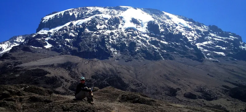 The glaciated summit dome of Kibo seen from the alpine saddle on Kilimanjaro, with a climber seated on rock for scale